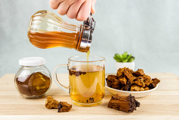Person makind tea drink with wild natural Chaga mushroom from birch tree, Inonotus obliquus pieces in tea glass on wooden table indoors home. Alternative traditional medicine. Pouring honey in tea.
