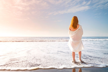 Redhead tender girl stand barefoot in the surf white foam on the beach. Woman in a white dress on the coast of an ocean, sunny morning