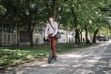 Young businessman with moustache driving scooter on sidewalk. Man in elegant clothes driving electric scooter on pavement with trees.