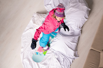 Above view of woman in warm clothes standing on snowboard at home with white duvet on floor like it...
