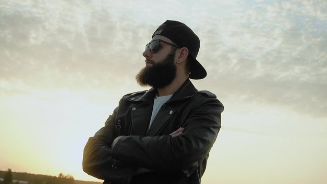 Bearded brutal image guy in black glasses, cap, leather jacket at sunset. Trendy fancy man stands still in slow motion. Fashionable stylish person with earring. Chic modish Life style. Biker. Clouds