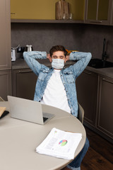 Fototapeta na wymiar Selective focus of freelancer in medical mask sitting at kitchen table with laptop and papers