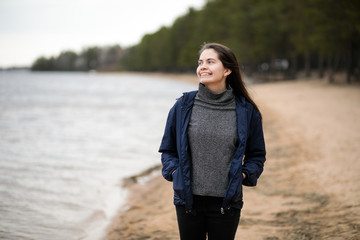 beautiful young girl stolt on the beach, yellow sand and forest, in a light jacket, smiling and looking to the side
