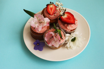 Homemade cupcakes decorated with fresh flowers and fresh strawberries on a white plate on a blue background
