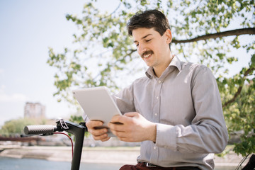 Male freelancer with moustache dressed with shirt holding tablet. Portrait of man in elegant outfit using tablet while sitting outdoor near scooter rudder.