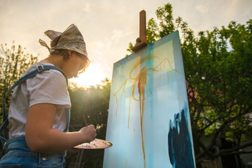 A young woman artist holds a brush and paints a picture on an easel in the rays of the sunset. The...