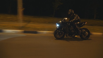 Man riding fast on modern black sport motorbike at evening city street. Motorcyclist racing his motorcycle on night empty road. Guy driving bike with headlight on. Concept of freedom. Side view