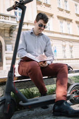 Smiling man in elegant outfit sitting on bench near scooter. Attractive guy wearing sunglasses sitting on bench and writing in notebook.