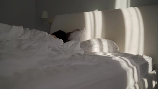 Woman sleeping in bed in the morning, dark brown long hair on pillow, sunlight falling on bed.