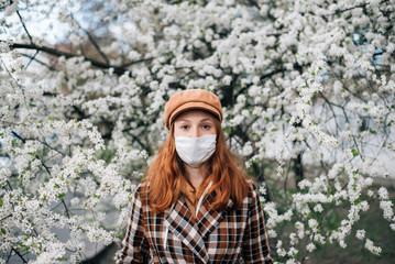 Fototapeta na wymiar redhead woman in a protective mask and a brown hat on a background of flowering trees. looking away