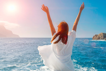 Happy redheadwoman wearing a white shirt breathing fresh air and raising arms on the beach of an...