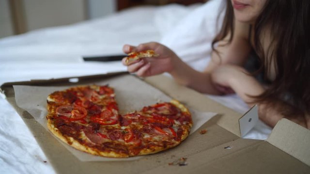 Woman eating pizza in bed, pizza served in a cardboard box, fast food delivery.