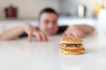 The man looks with cunning eyes at a cheeseburger lying on the table. Burger is not healthy food....
