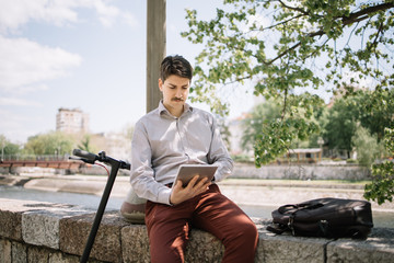 Obraz premium Guy with moustache sitting on stone fence near river. Man in elegant shirt using tablet while sitting on river fence near scooter.