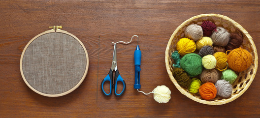 Colored woolen and acrylic balls of yarn in wicker basket, special multi-level needle for embroidery using the technique of punching tapestry or carpet embroidery and a canvas in hoop on wooden table