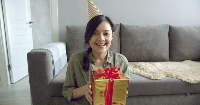 Happy young woman in a birthday cap holding a gift recording a video or chatting online with his friends at home. Vlogging, social networks and online communication.