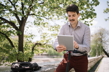 Obraz premium Male freelancer on scooter working on tablet outdoor. Boy with moustache using tablet while leaning on scooter rudder with tree in the background.