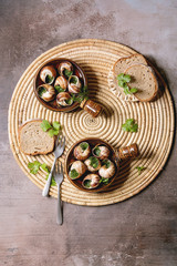 Escargots de Bourgogne - Snails with herbs butter, gourmet dish, in two traditional ceramic pans with parsley and bread on straw napkin over brown textured background. Flat lay, space