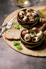 Escargots de Bourgogne - Snails with herbs butter, gourmet dish, in two traditional ceramic pans with coriander, bread and glass of white wine on straw napkin over brown textured background.