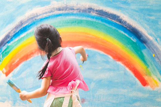 Asian little girl is painting the colorful rainbow and sky on the wall and she look happy and funny, concept of art education and learn through play activity for kid development.