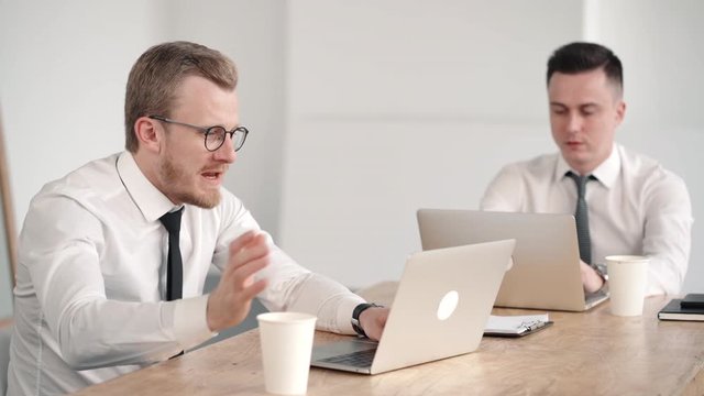 Two young male coworkers working on laptops and discussing business. Two successful businessmen investor wear shirt and drink coffee. talking communicating about proccess