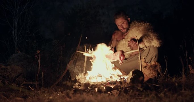 Wild bearded man making cuts on frying animal body and evading hot fire then starting to sharpen knife at night in wilderness.