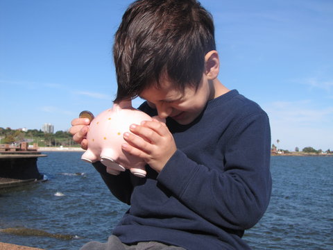 Money saving in piggy bank. A young boy looks inside a pink ceramic piggy bank before putting a coin in the waterfront with the sea in the background illuminated by sunlight.