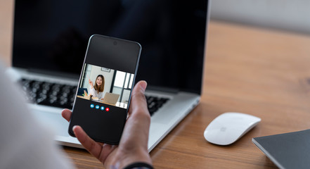 Young woman holding smartphone and making video call with her friend while sitting at office desk.