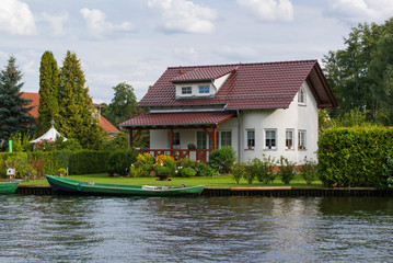 Beautiful white house with red roof on the river bank. In front of it there is a green lawn and a...