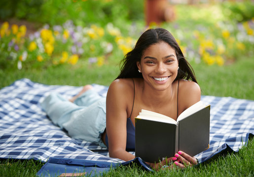 Cute Young African American Woman Reading Book On Picnic Blanket In Shade - In Front Of Blooming Flowers