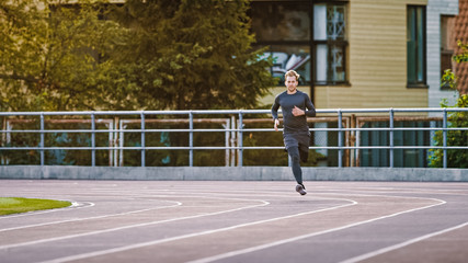 Smiling Athletic Fit Man in Grey Shirt and Shorts Jogging in the Stadium. He is Running Fast on a Warm Summer Afternoon. Athlete Doing His Routine Sports Practice.