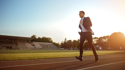 Young Serious Businessman in a Suit Running in an Outdoors Stadium. He Wears Glasses and is Holding...