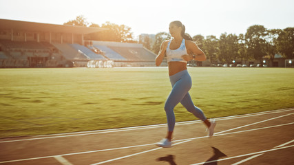 Fototapeta premium Beautiful Fitness Girl in Light Blue Athletic Top and Leggings Jogging in the Stadium. She is Running on a Warm Summer Afternoon. Athlete Doing Her Routine Sports Practice on a Track.
