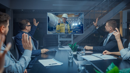 Obraz na płótnie Canvas Late at Night In Corporate Meeting Room: Executives and Businesspeople Sitting at Negotiations Table Having Video Conference Call with Professional Female Industrial Engineer. They Do Hello Gesture.