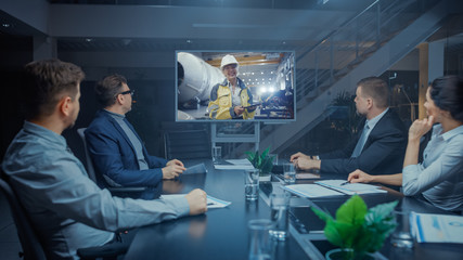 Late at Night In Corporate Meeting Room: Executives and Businesspeople Sitting at Negotiations Table Having Video Conference Call with Professional Female Industrial Engineer in Factory.