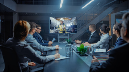 Late at Night In Corporate Meeting Room: Executives and Businesspeople Sitting at Negotiations Table Having Video Conference Call with Professional Female Industrial Engineer in Factory.