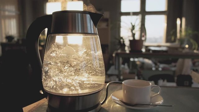 A transparent kettle of water boils against the background of the sunset shining through the window. The concept of coffee break and end of the working day.