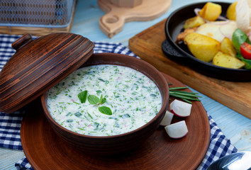 Okroshka - summer cold soup. Cold soup with fresh cucumbers, radishes, potato and sausage. Traditional russian food. Okroshka with kefir and rosemary in a clay plate with a lid.
