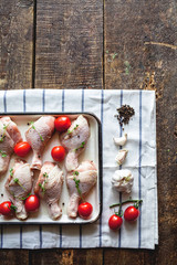Raw chicken legs in a pan for baking with herbs and tomatoes on a tablecloth on a wooden table. View from above. Preparation of dinner. Dietary food