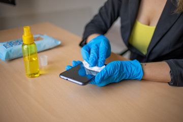 Woman's hand in blue gloves sanitizing cleaning smartphone mobile phone with alcohol on wood table surface with wet wipes
