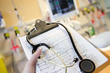 An ECG (electrocardiogram) is featured with a black stethoscope in a hospital room with a monitor in the back ground.