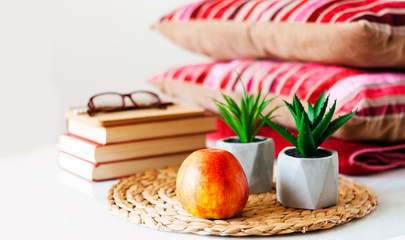 Cozy home interior decor: apple, stack of books, plants in pots on a wicker stand, pillows and plaid on a white table. Distance home education. Quarantine concept of stay home.