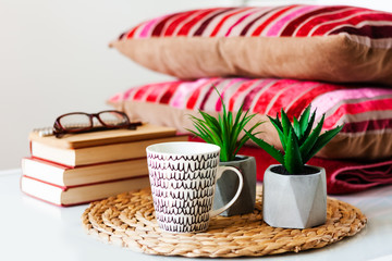 Cozy home interior decor: cup of coffee, stack of books, plants in pots on a wicker stand, pillows and plaid on a white table. Distance home education. Quarantine concept of stay home.