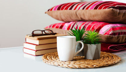 Cozy home interior decor: cup of coffee, stack of books, plants in pots on a wicker stand, pillows and plaid on a white table. Distance home education. Quarantine concept of stay home.