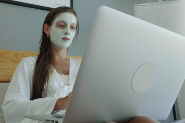 Close up of thoughtful concerned woman with cosmetic facial mask working on laptop computer lying in the bed 