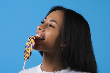 Close-up of fashionable gorgeous african american woman with short straight black hair enjoying sweet lollipop, smiling and looking at camera on blue background