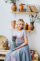 Blonde young woman holding a flower and metal watering can in the interior of an eco clay pot and wooden pallets. DIY hand made interior concept