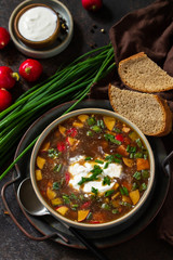 Traditional dish Russian cuisine. Cold summer Okroshka soup with kvass, sausage and vegetables in a bowl on a dark stone countertop.