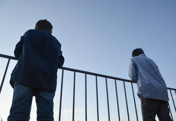 Two children on their backs in full social distancing by the covid-19 on a background of blue sky and leaning on an iron railing looking at the horizon. Stay home to prevent the coronavirus pandemic. 