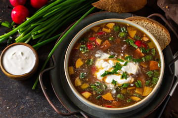 Traditional dish Russian cuisine. Cold summer Okroshka soup with kvass, sausage and vegetables in a bowl on a dark stone countertop.